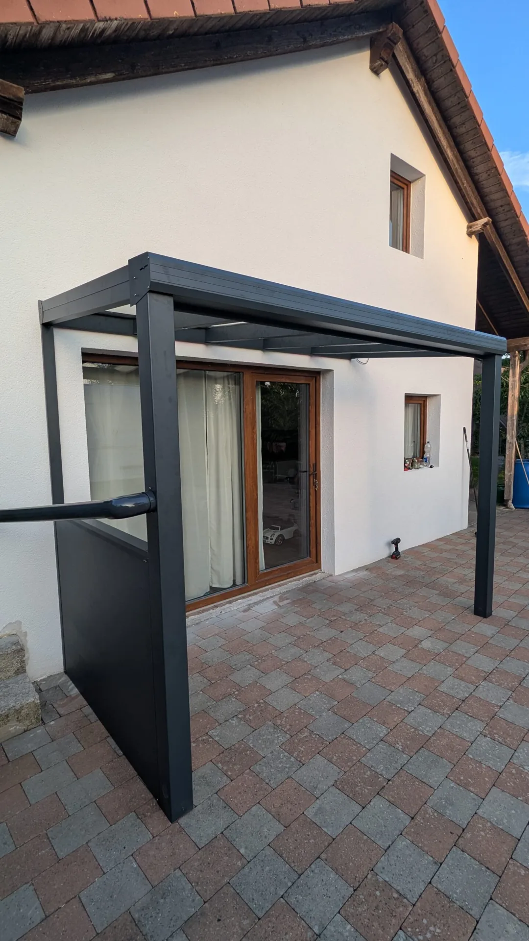 Anthrazitfarbene Dachkonstruktion mit einer seitlichen Sichtschutzwand am gepflasterten Sitzplatz.